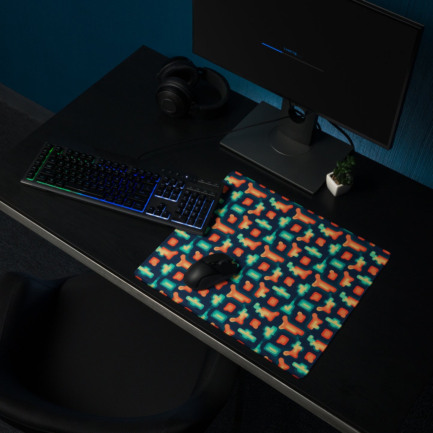 An 18" x 16" gaming desk pad with blue and orange puzzle pieces on a dark blue background. It sits on a black desk with a keyboard, monitor, and mouse.