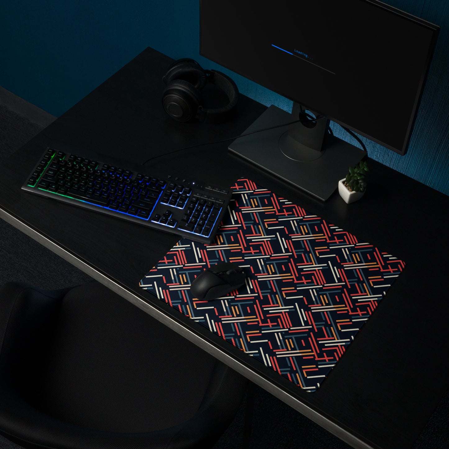An 18" x 16" gaming desk pad with red, blue, white, and orange lines on a black background. It sits on a black desk with a monitor, keyboard, and mouse.