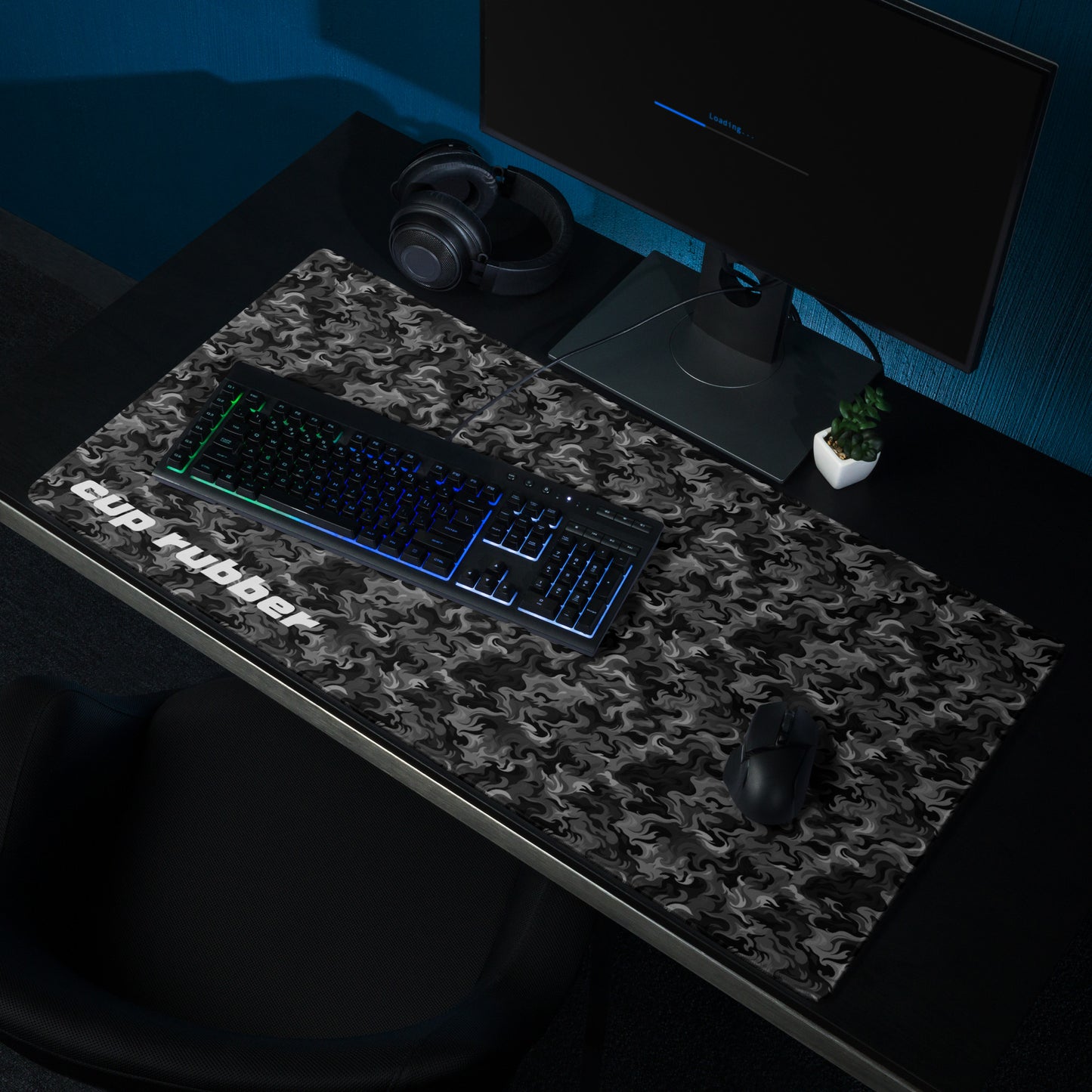 A 36" x 18" desk pad with a camo pattern all over it and the word "Cup Rubber" in the bottom left corner shown on a desk setup. Black and Charcoal in color.
