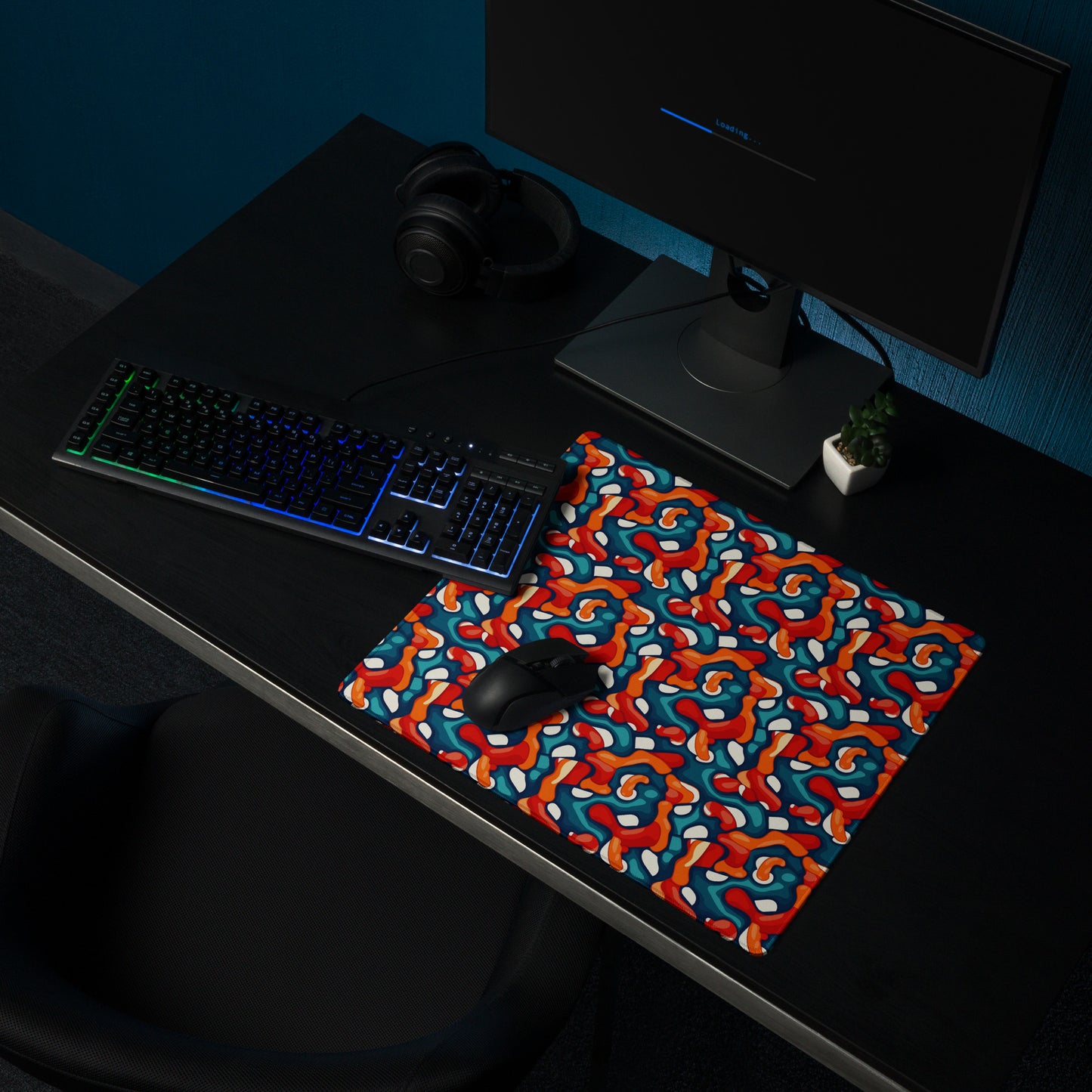 A 18" x 16" desk pad with abstract line art on it shown on a desk setup. Red, Teal and Orange in color.