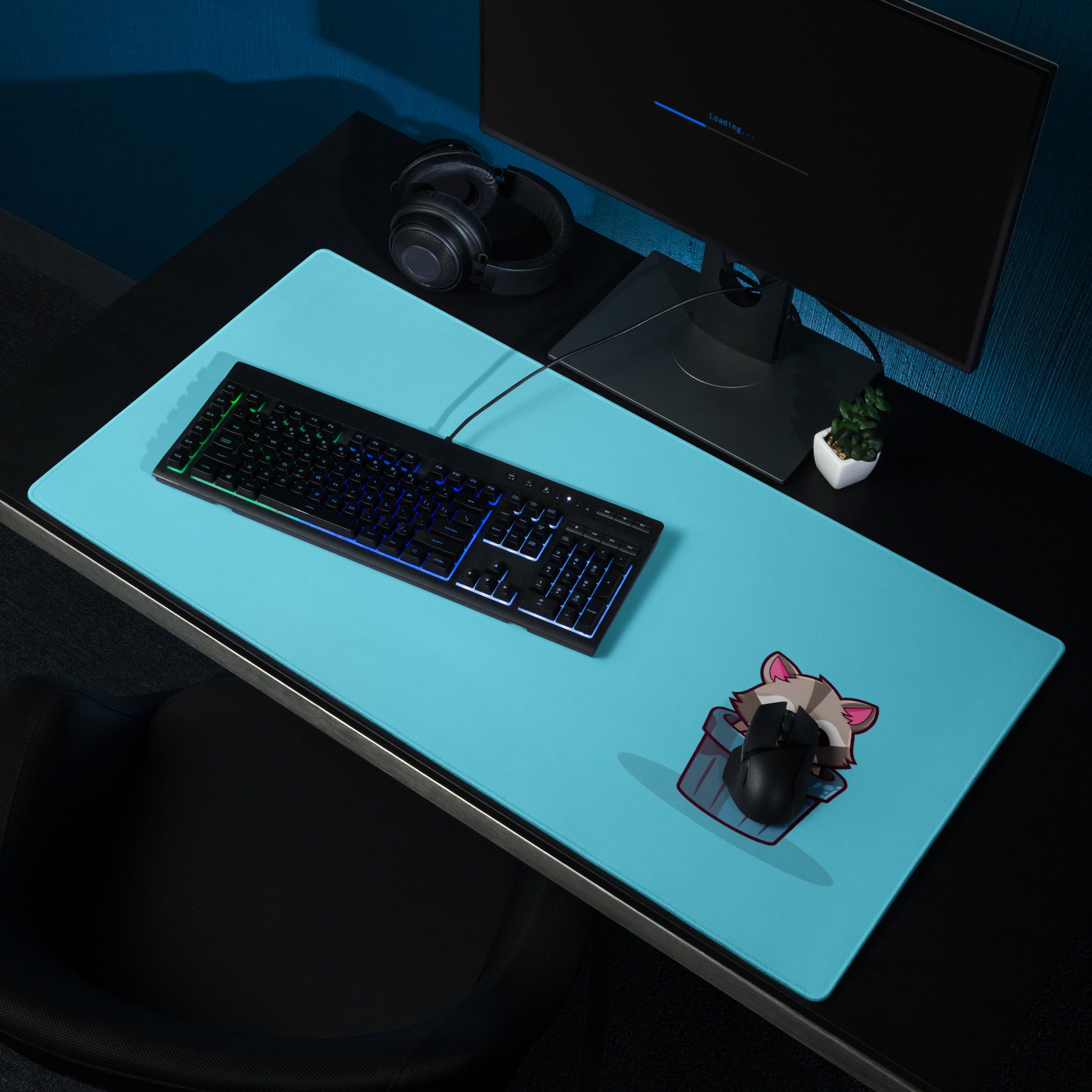A gaming desk pad with a kawaii raccoon in a garbage can on a blue background. A keyboard and mouse sit on it.