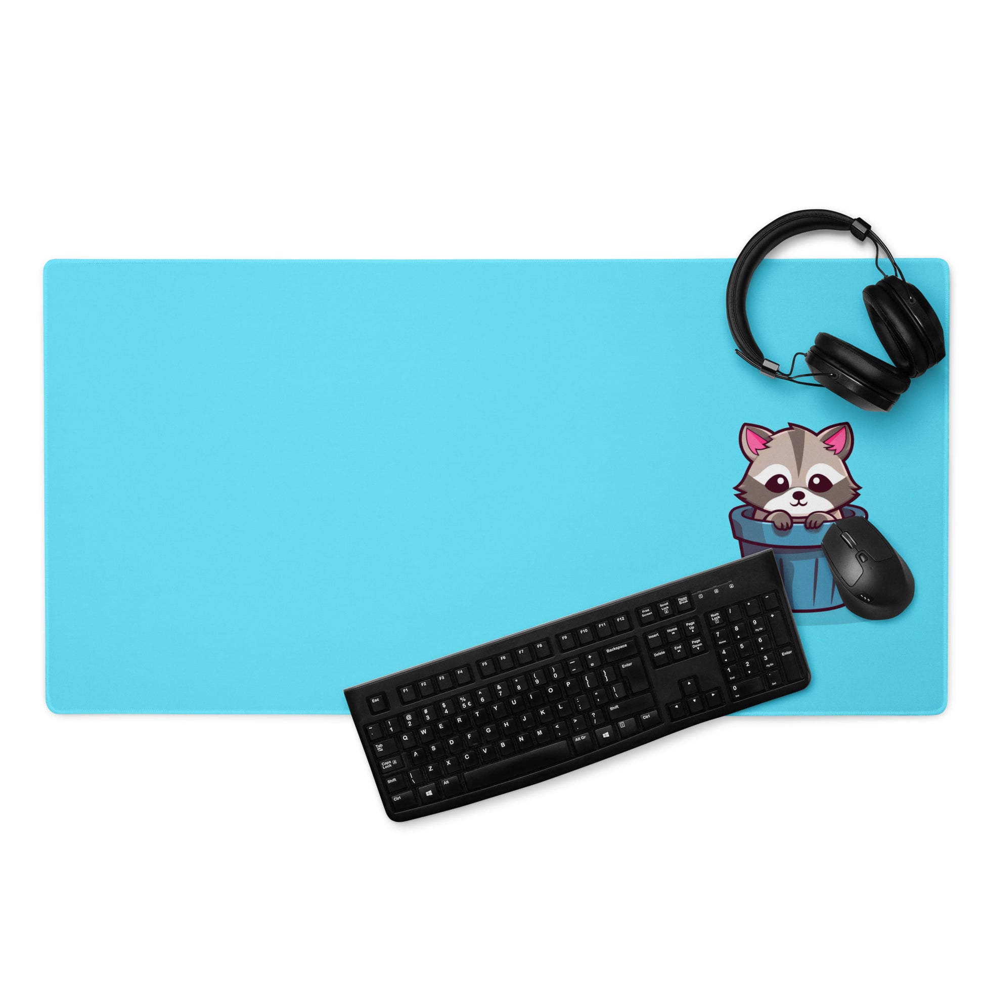 A gaming desk pad with a kawaii raccoon in a garbage can on a blue background. A keyboard, mouse, and headphones sit on it.