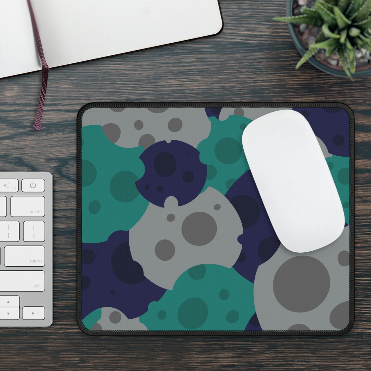 A gaming mouse pad with gray, teal, and dark blue meteorites sitting on a desk. A mouse sits on top of it.
