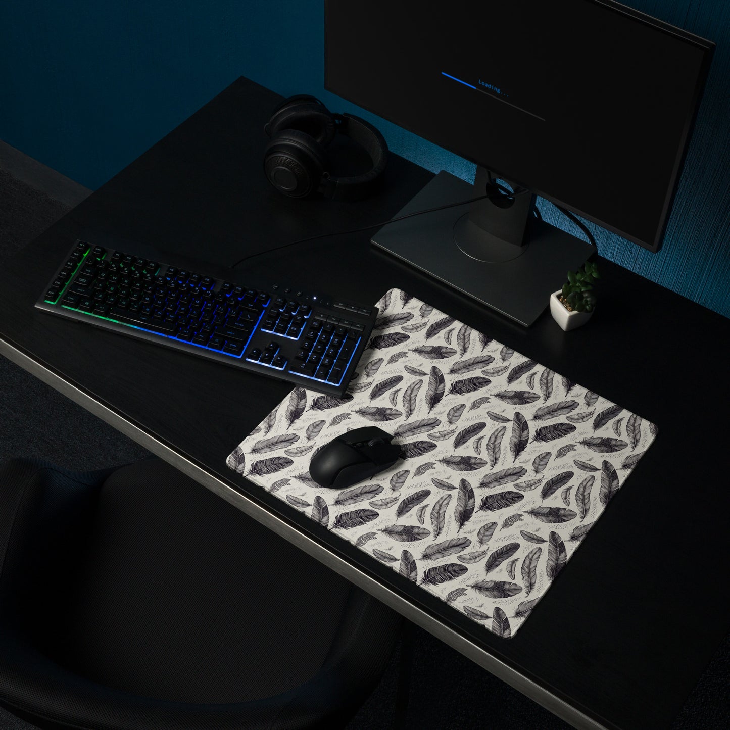 An 18" x 16" gaming desk pad with black and white feathers. It sits on a black desk with a keyboard, mouse, and monitor.