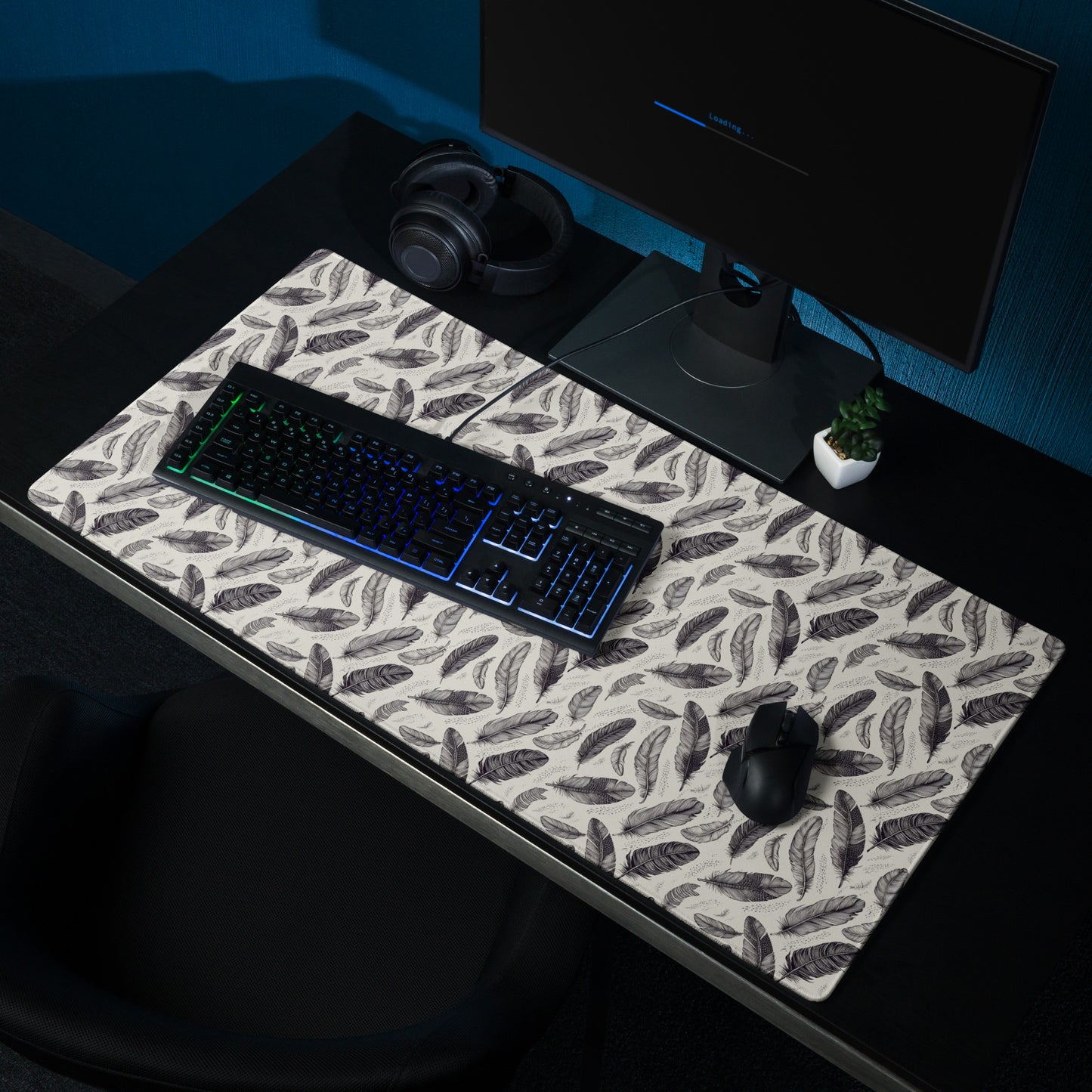 A 36" x 18" gaming desk pad with black and white feathers. It sits on a black desk with a keyboard, mouse, and monitor.