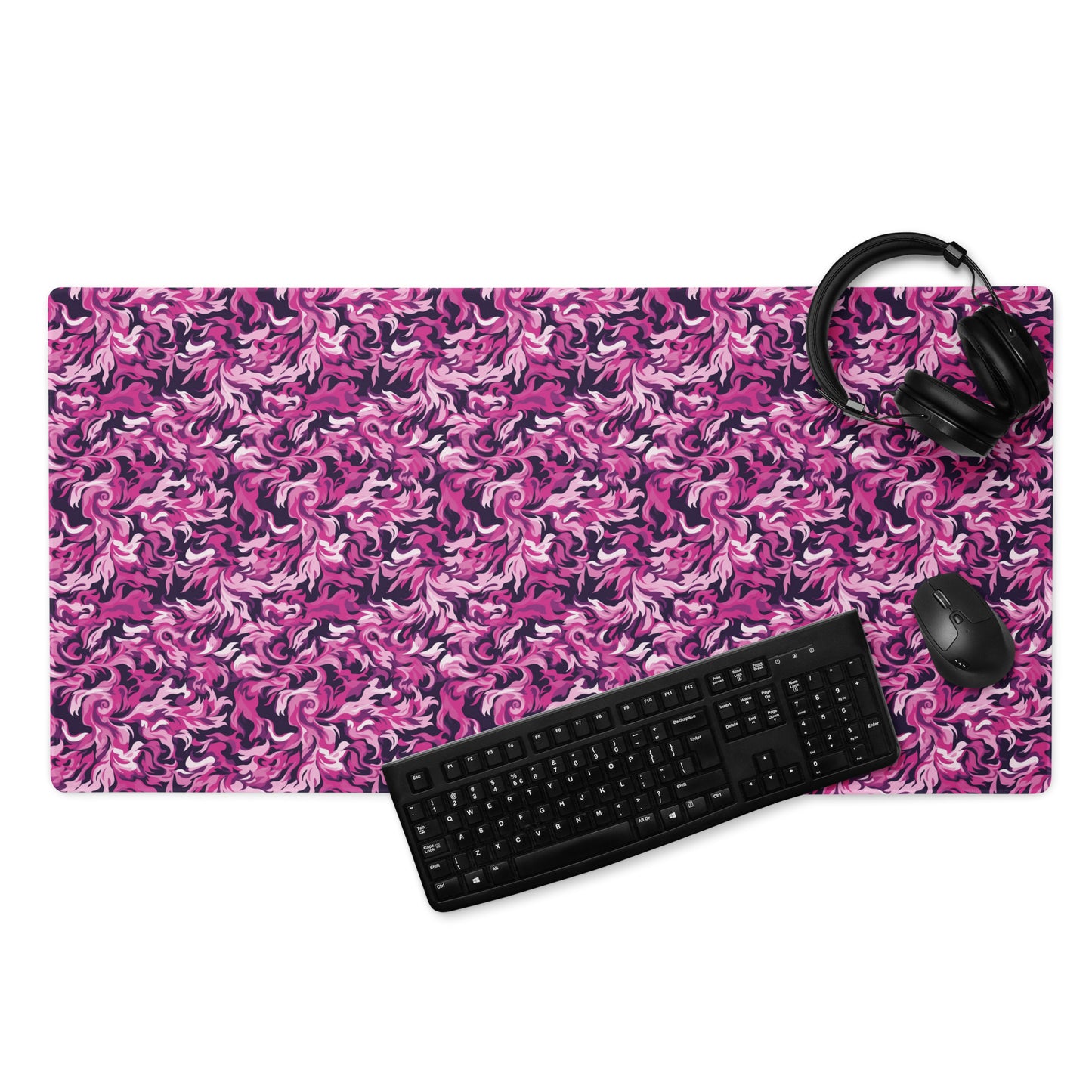 A 36" x 18" desk pad with a pink and magenta camo pattern. With a keyboard, mouse, and headphones sitting on it.