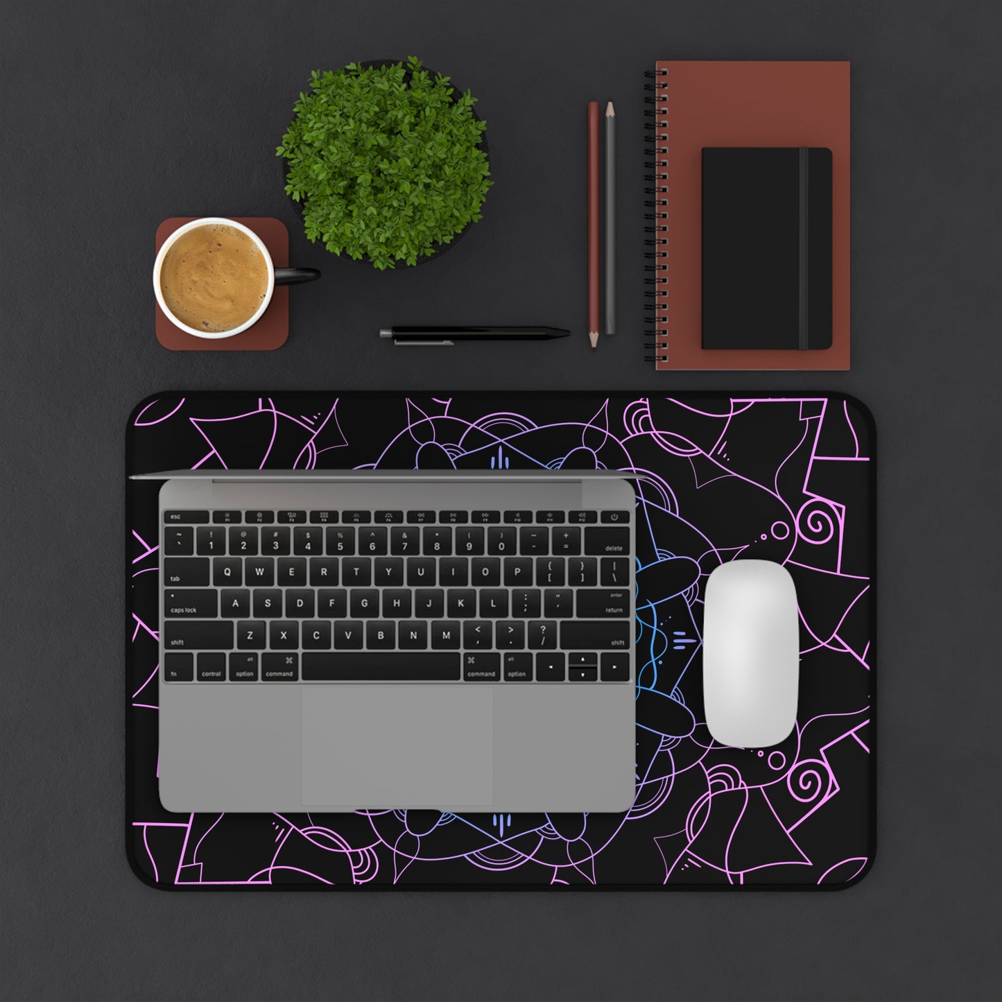 A 12" x 18" desk mat with a pink and blue mandala pattern on a black background. A laptop and mouse sit on top of it.