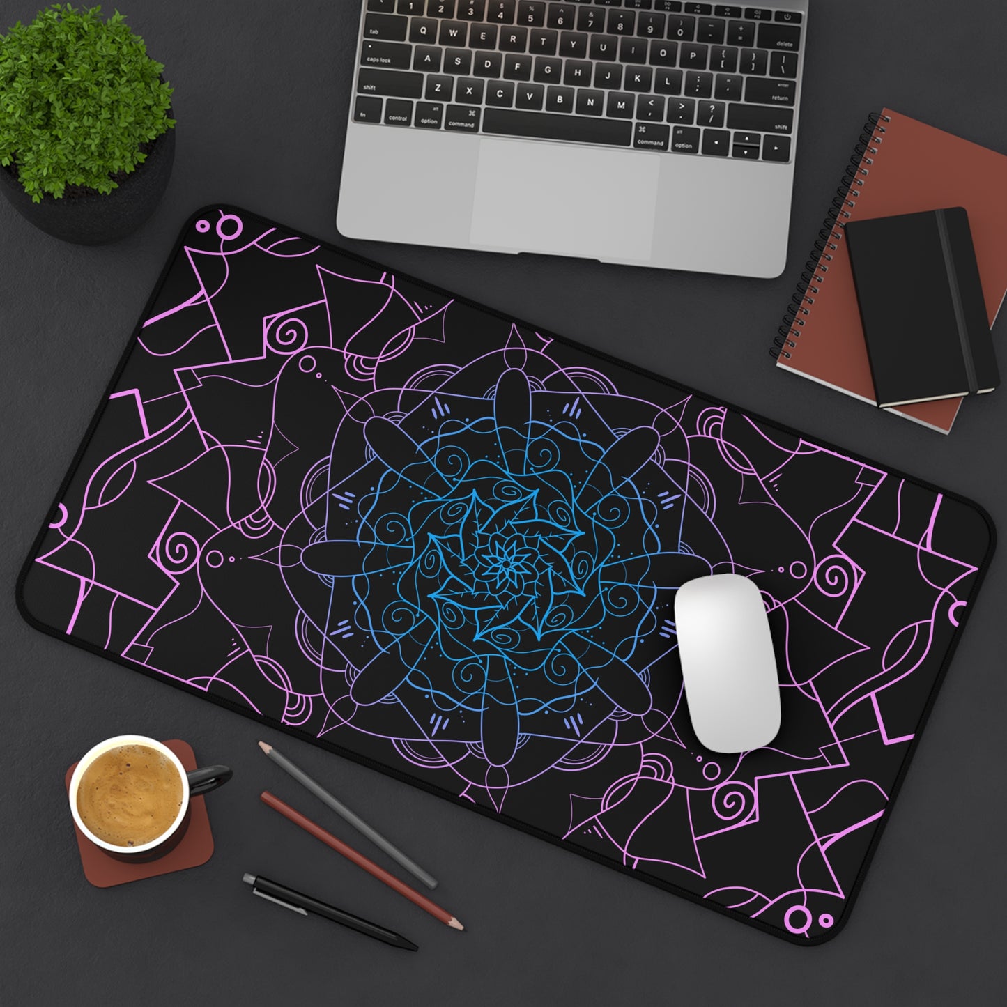 A 12" x 22" desk mat with a pink and blue mandala pattern on a black background sitting at an angle.