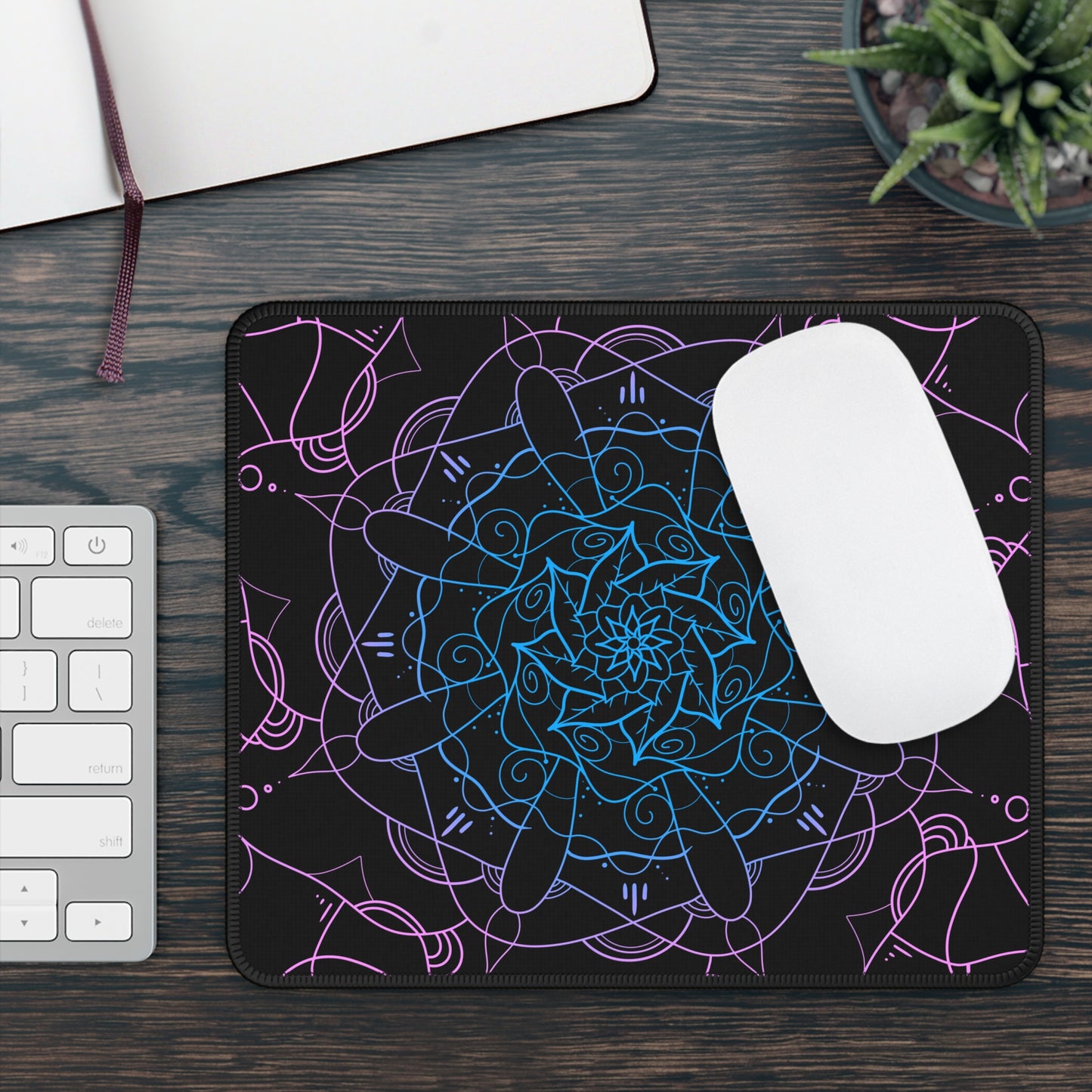 A gaming mouse pad with a pink and blue mandala pattern on a black background. A mouse sits on top of it.