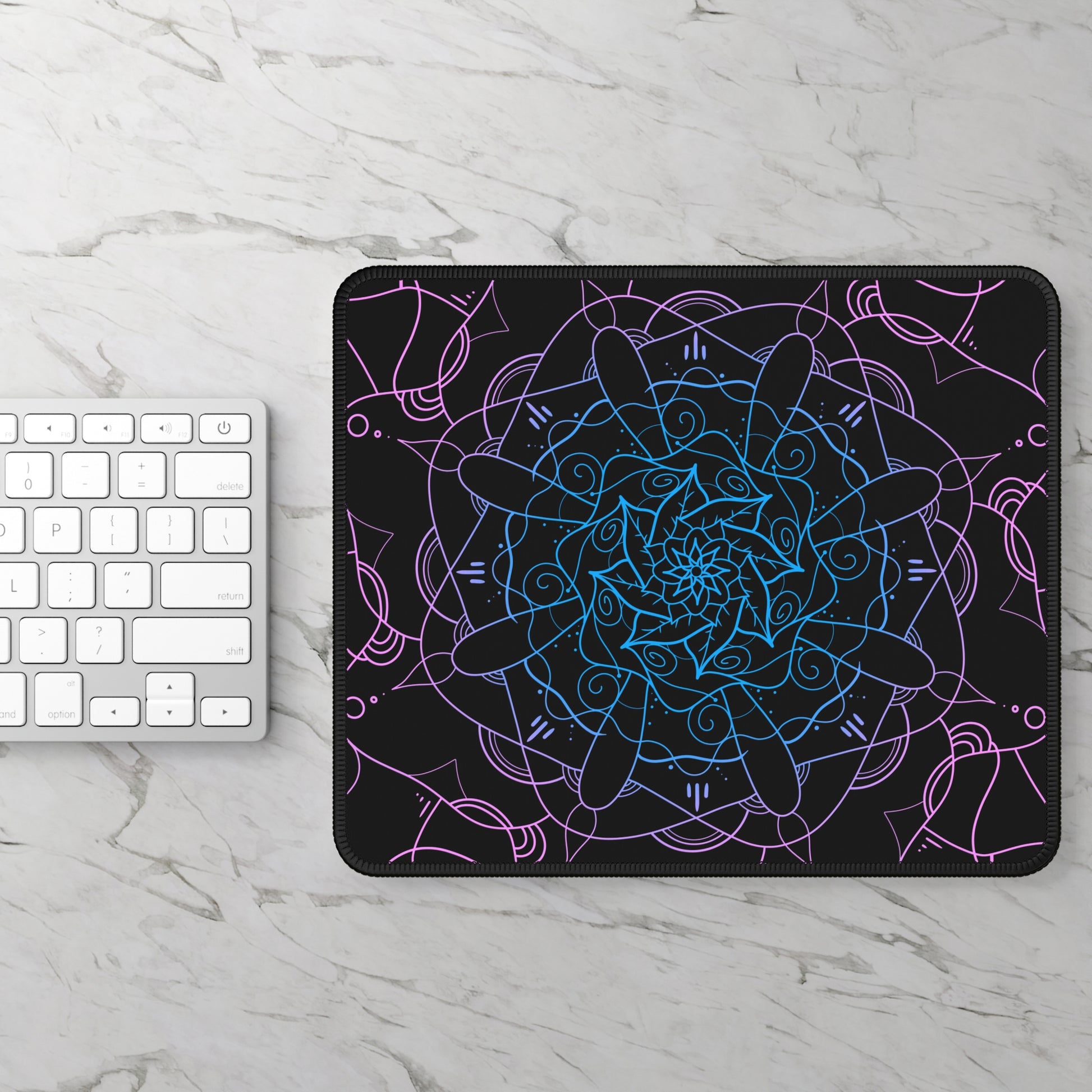 A gaming mouse pad with a pink and blue mandala pattern on a black background sitting next to a keyboard.
