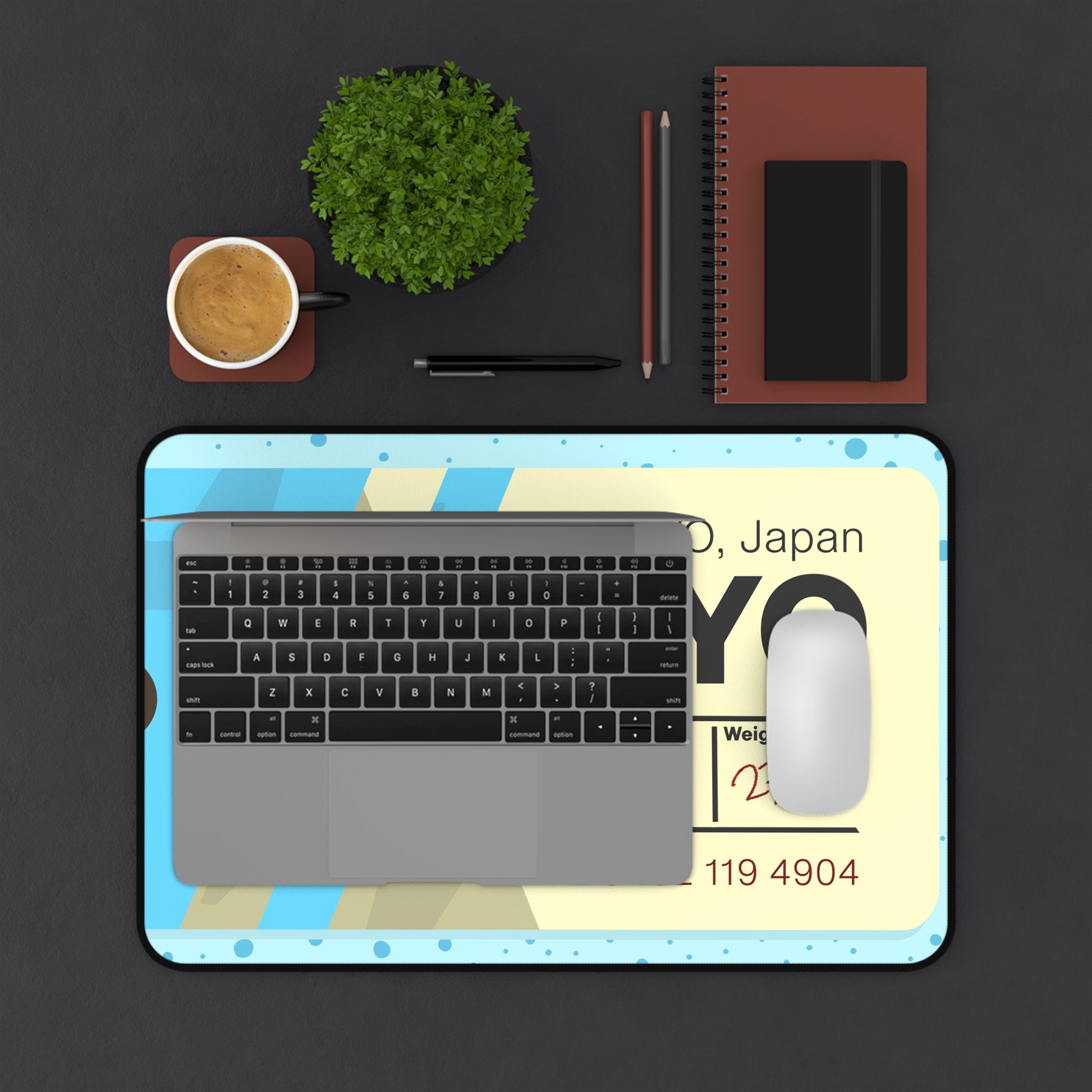 A 12" x 18" desk mat with a dotted blue background and a blue and yellow Tokyo airline baggage tag on it. A laptop and mouse sit on top of it.