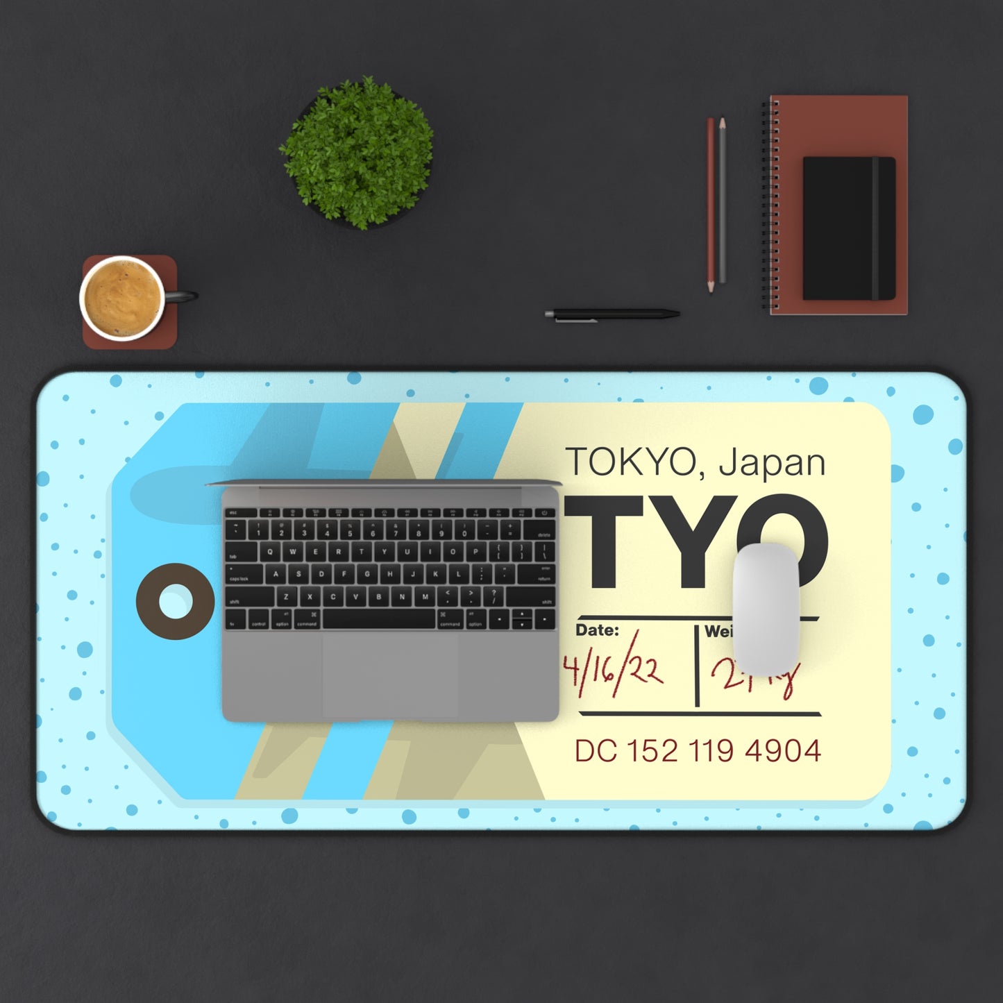 A 31" x 15.5" desk mat with a dotted blue background and a blue and yellow Tokyo airline baggage tag on it. A laptop and mouse sit on top of it.