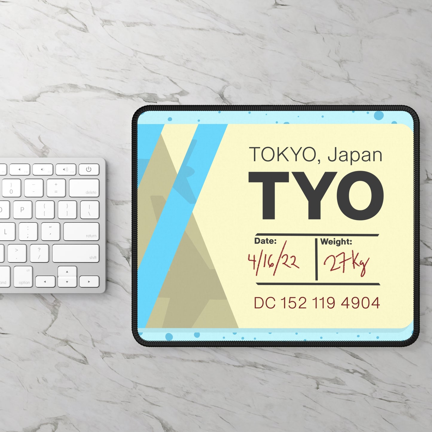 A gaming mouse pad with a dotted blue background and a blue and yellow Tokyo airline baggage tag on it sitting next to a keyboard.