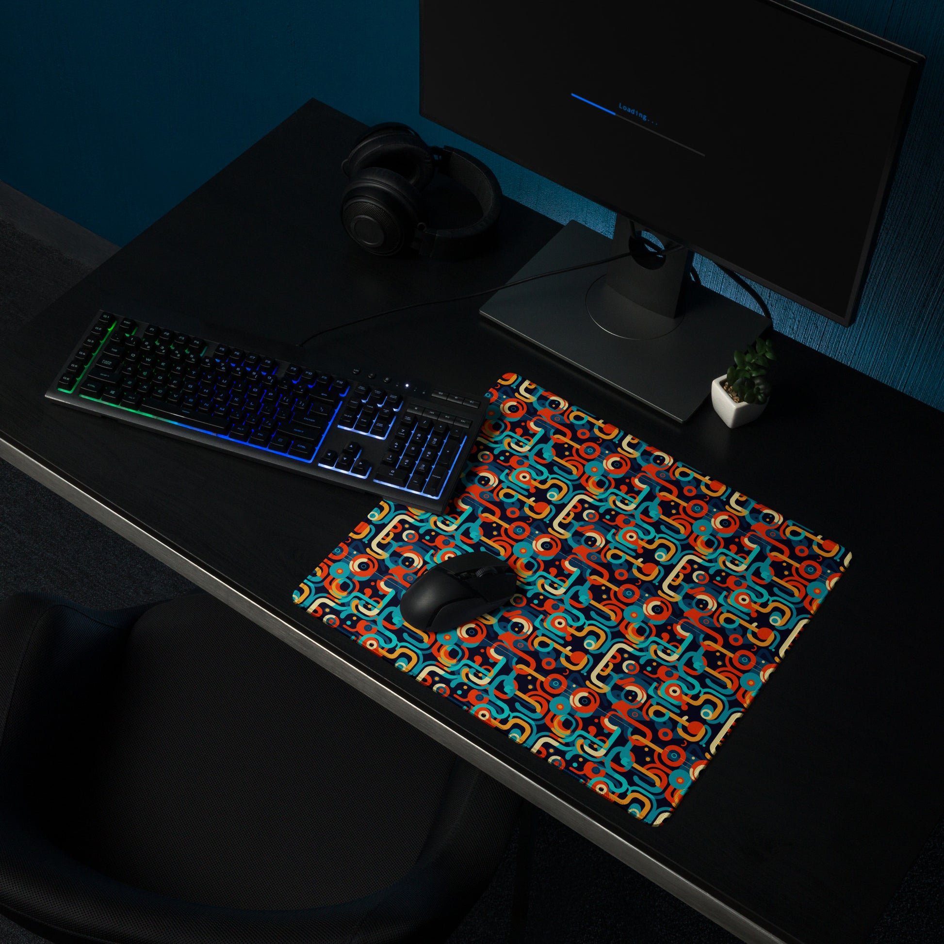 An 18" x 16" gaming desk pad with blue, orange, and beige lines on a black background. It sits on a black desk with a keyboard, monitor, and mouse.