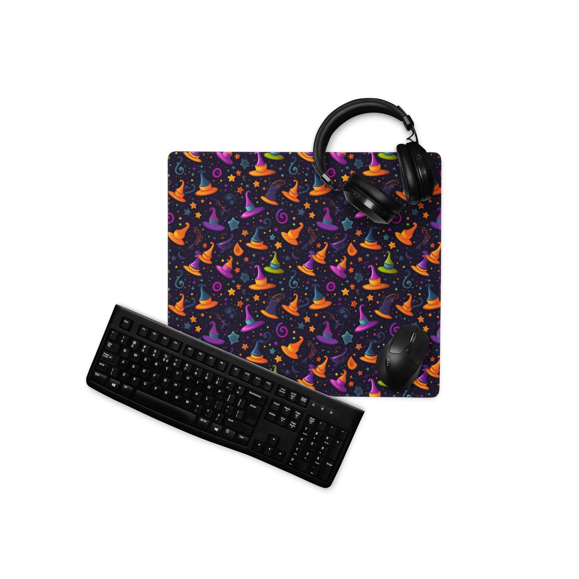 A 18" x 16" desk pad with a orange and purple wizard hat pattern. With a keyboard, mouse, and headphones sitting on it.