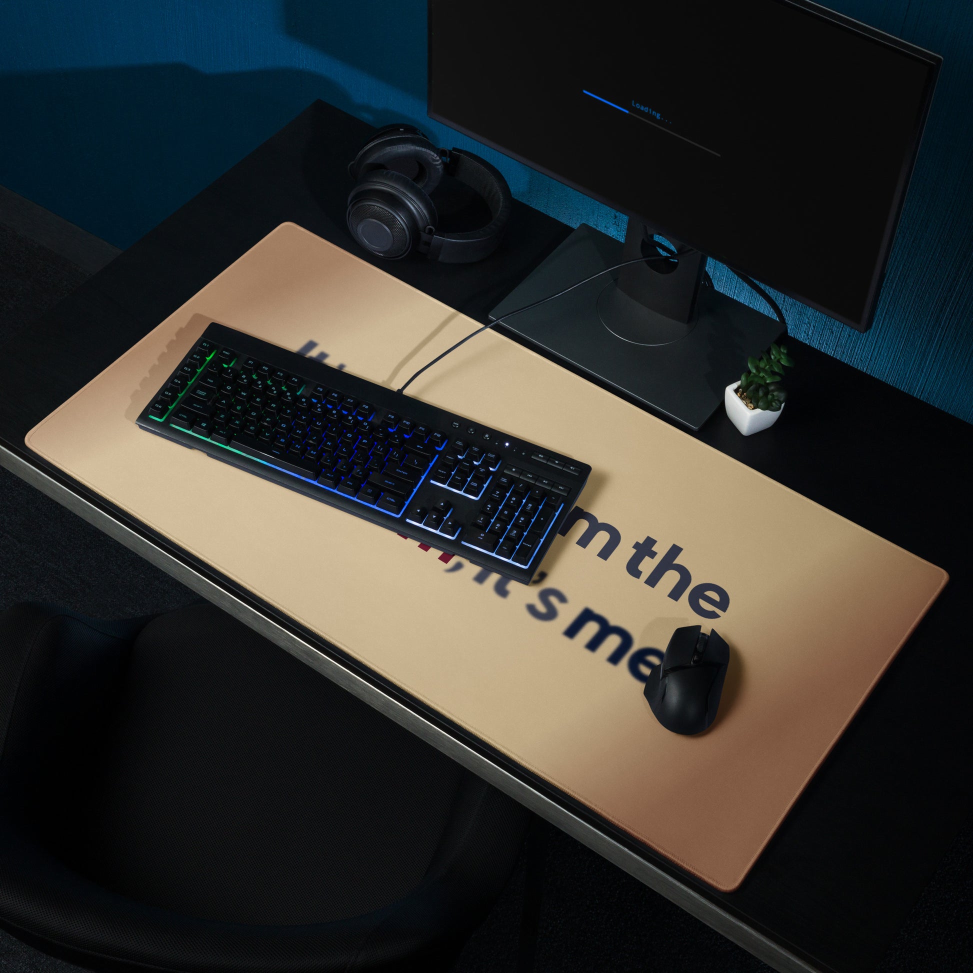 A beige gaming desk pad with the quote It's me, hi! I'm the problem, it's me. The desk pad sits on a black desk with a monitor, keyboard, and mouse.