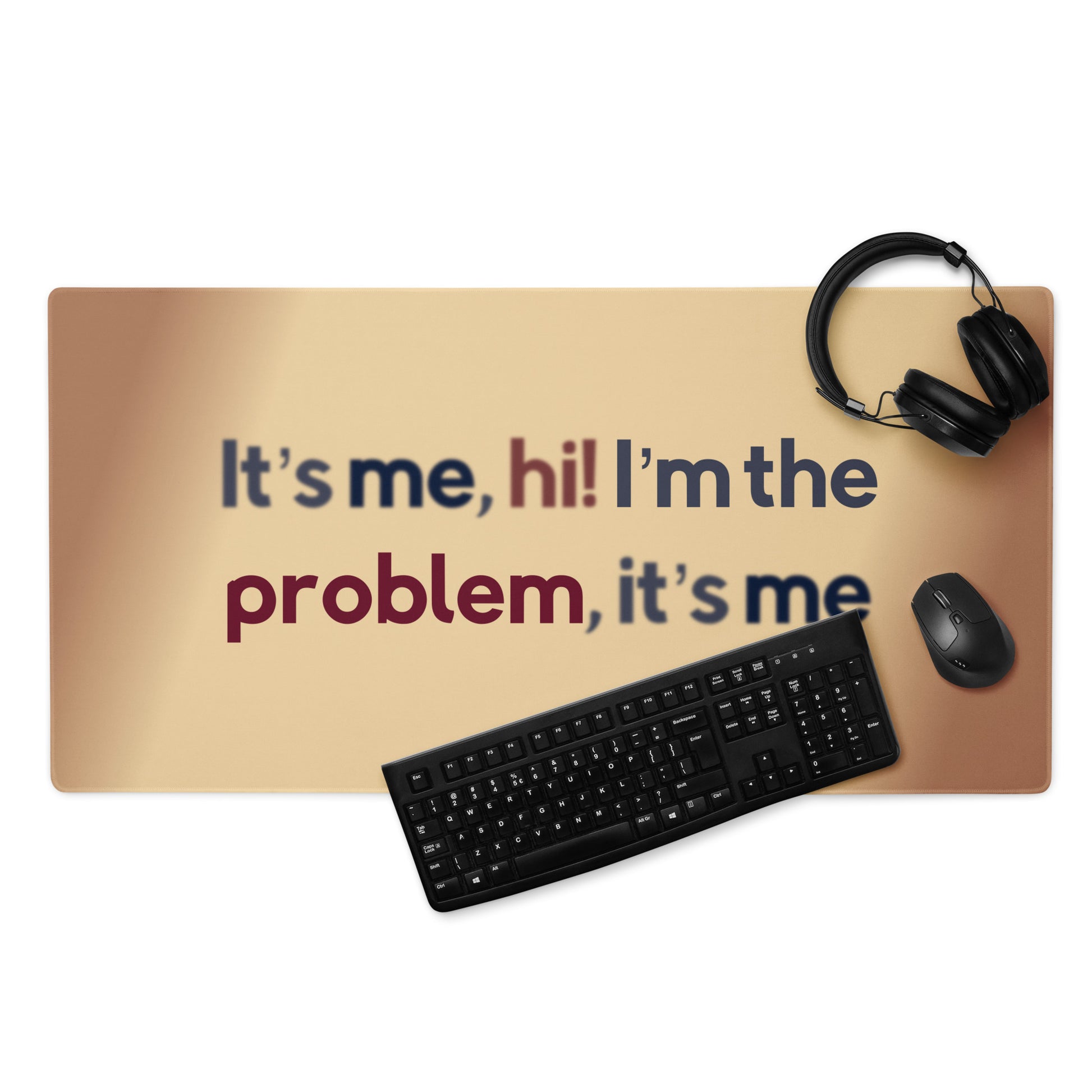 A beige gaming desk pad with the quote It's me, hi! I'm the problem, it's me. A keyboard, headphones, and mouse sit on the desk pad.