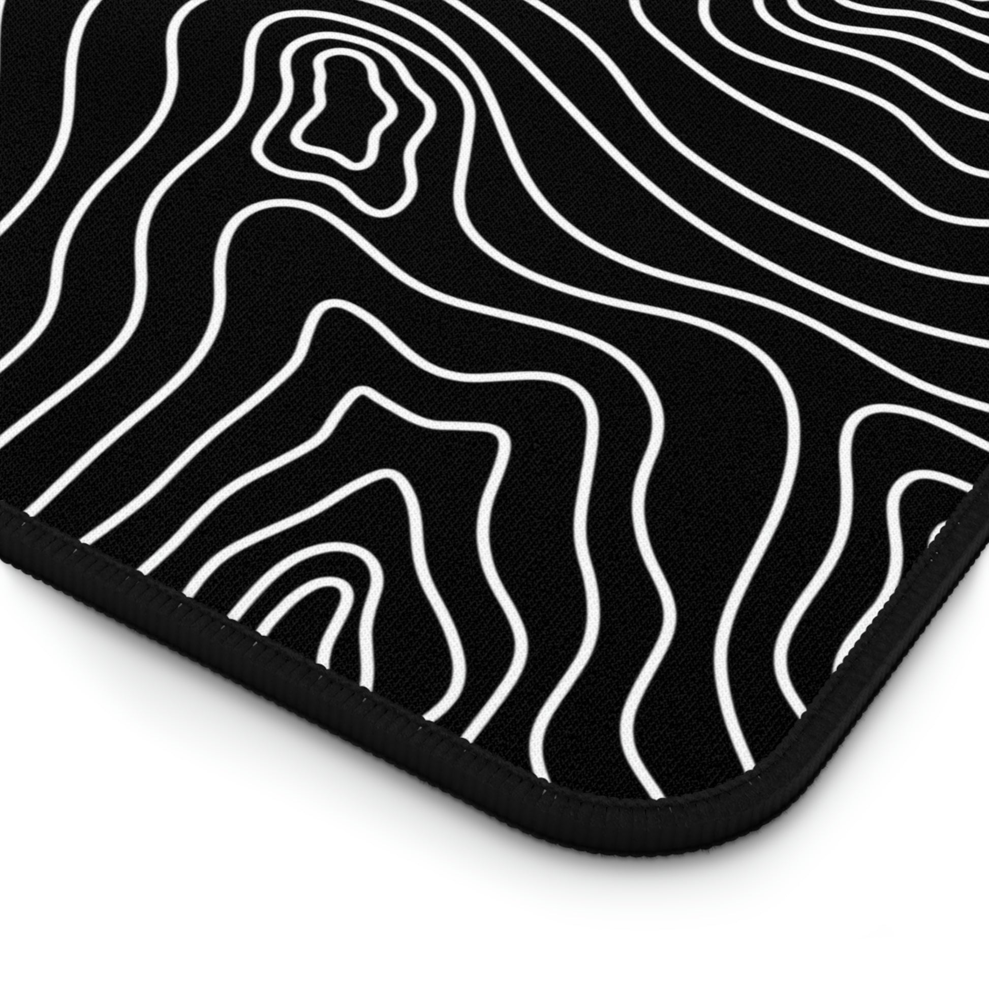The bottom right corner of a 12" x 18" black desk mat with white topographic lines.