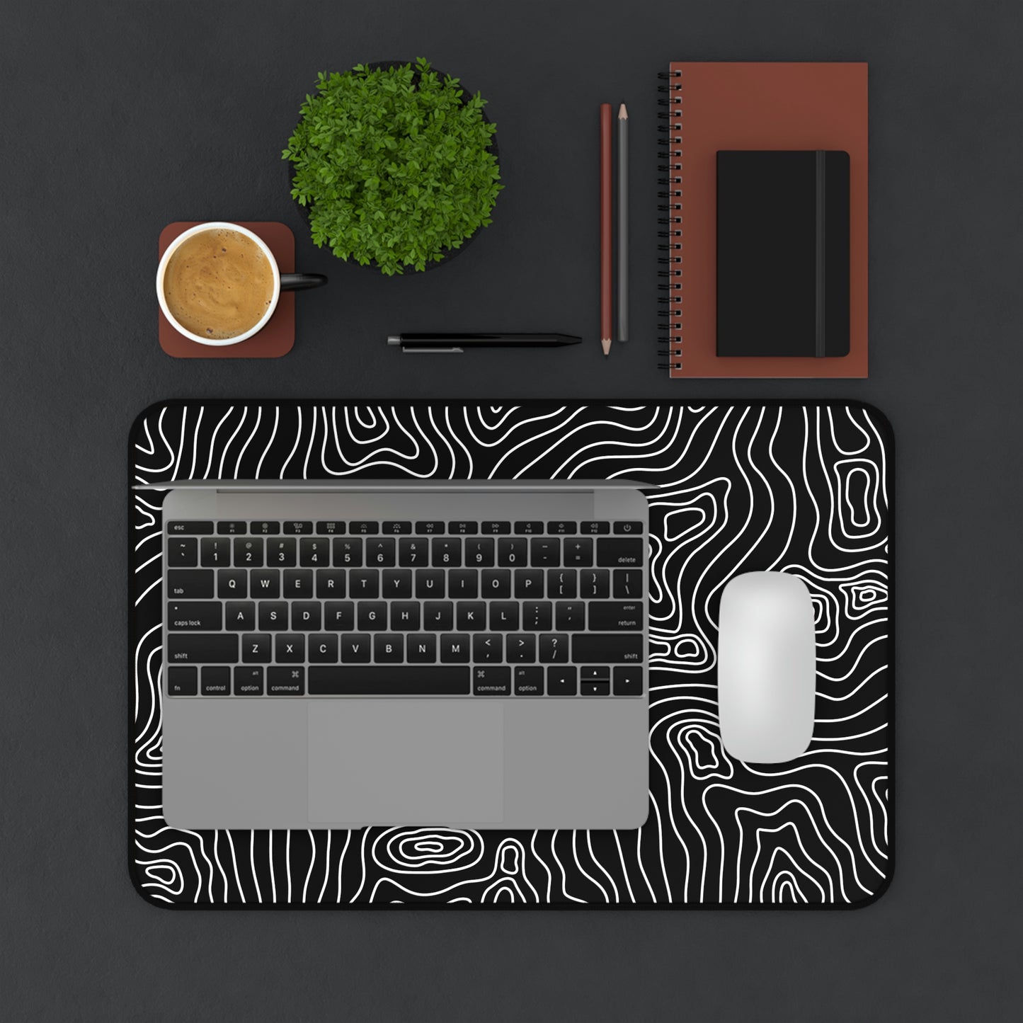 A 12" x 18" black desk mat with white topographic lines. A laptop and mouse sit on top of it.