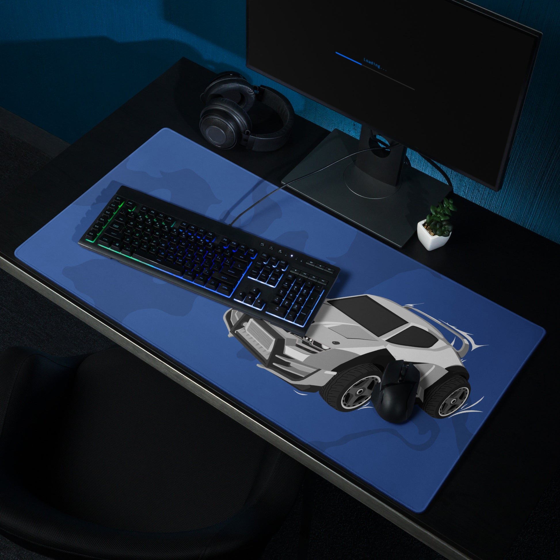 A blue gaming desk pad with a gray Takumi. The desk pad sits on a black desk with a monitor, keyboard, and mouse.