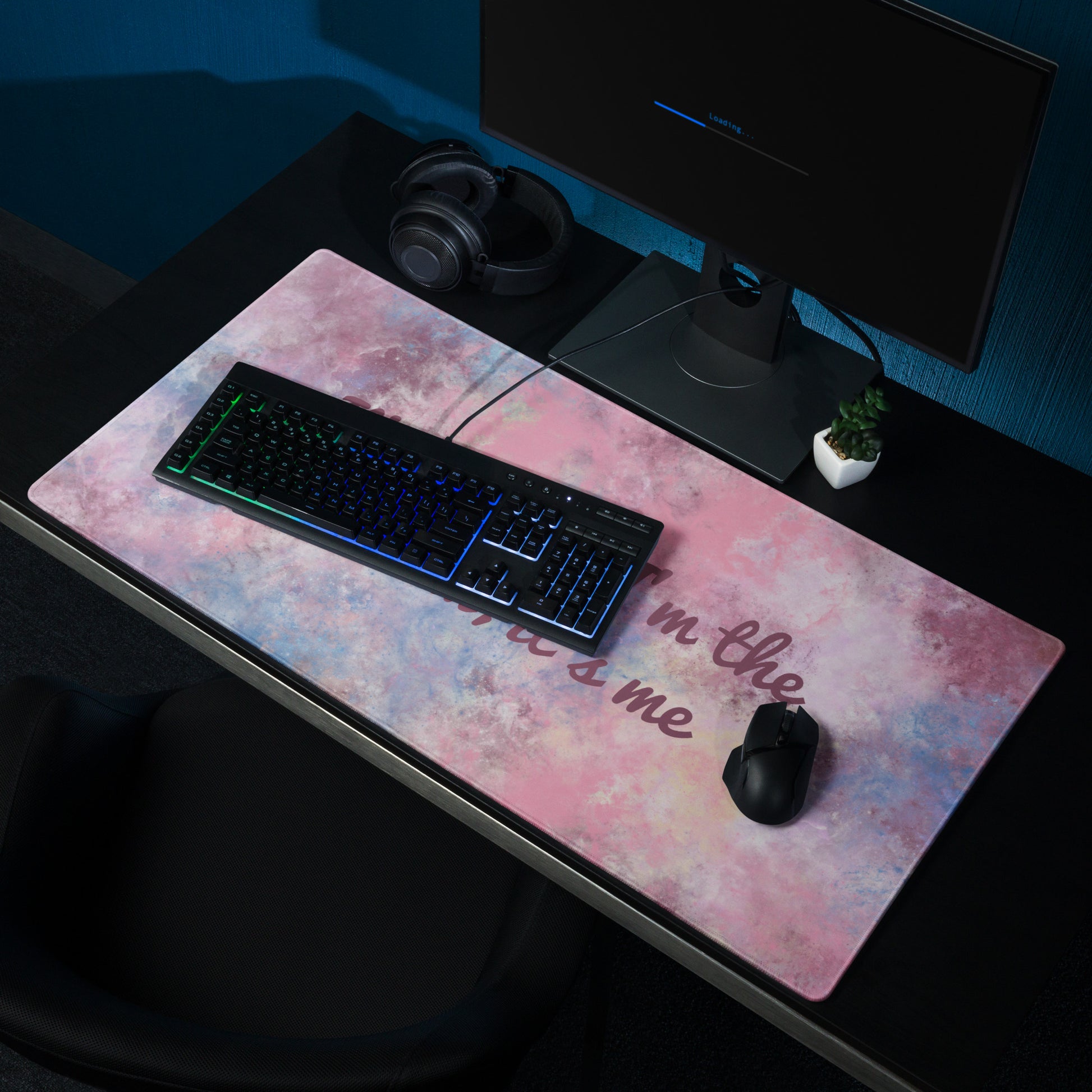 A pink, blue, and white gaming desk pad with the quote It's me, hi! I'm the problem, it's me. The desk pad sits on a black desk with a monitor, keyboard, and mouse.