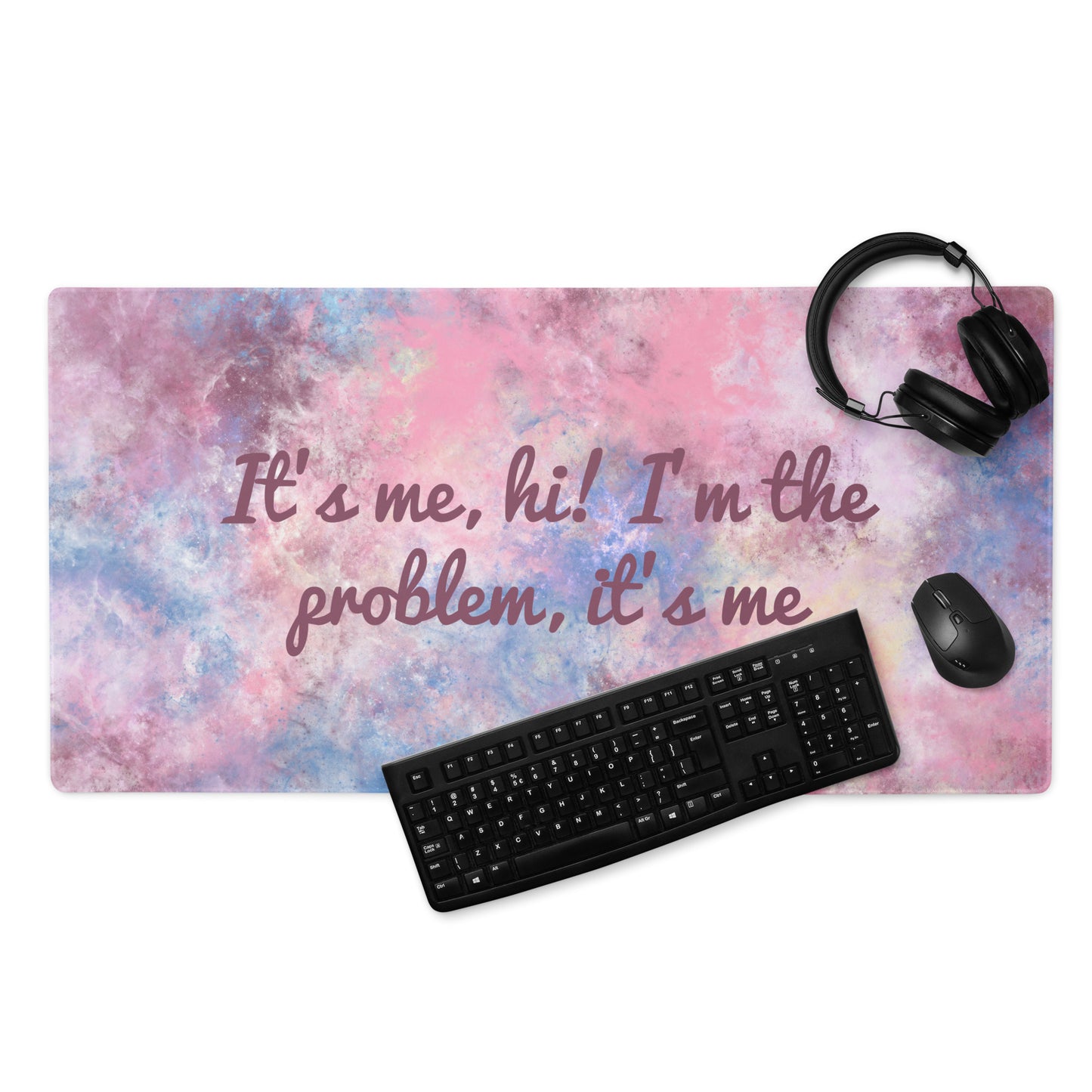 A pink, blue, and white gaming desk pad with the quote It's me, hi! I'm the problem, it's me. A keyboard, headphones, and mouse sit on the desk pad.
