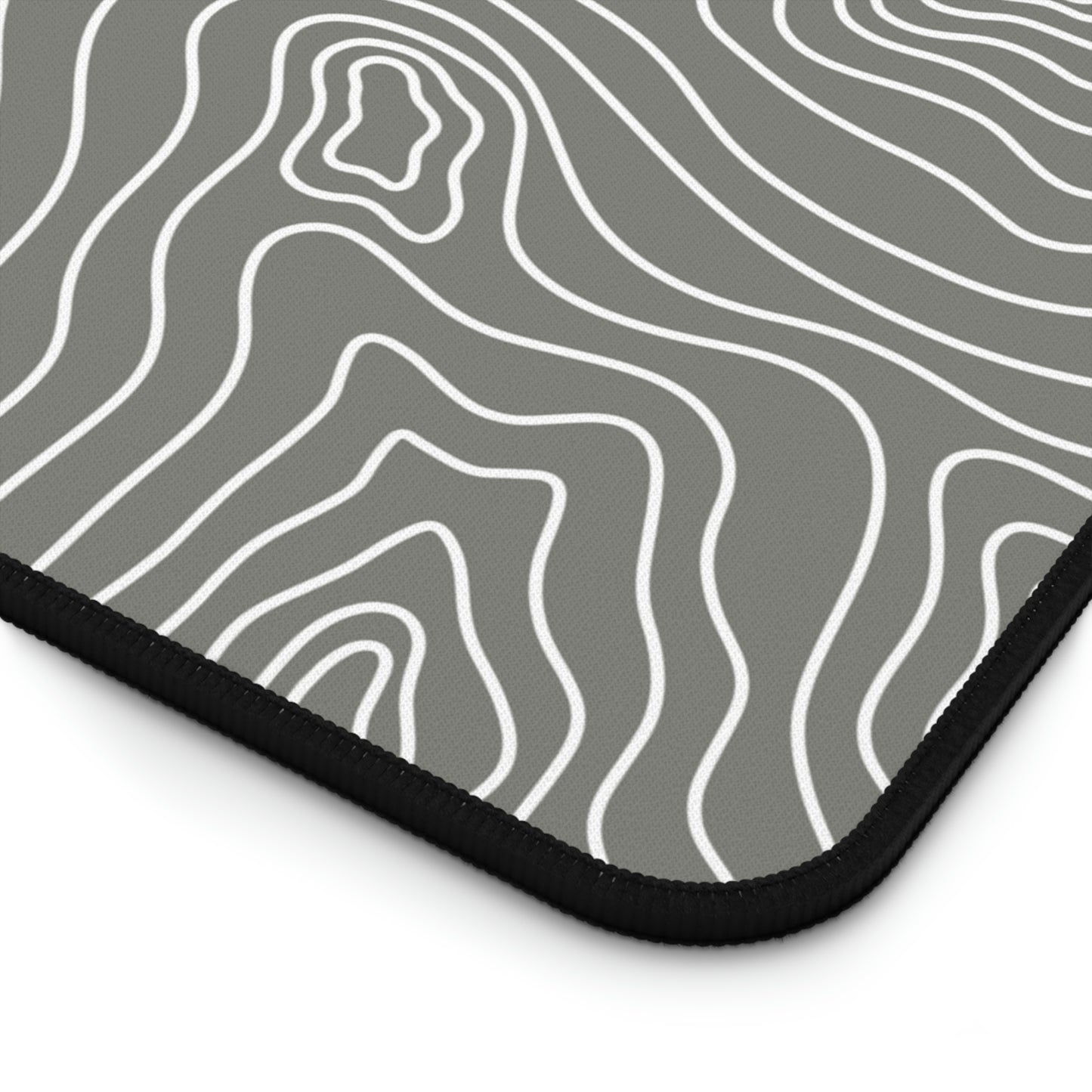 The bottom right corner of a 12" x 18" gray desk mat with white topographic lines.