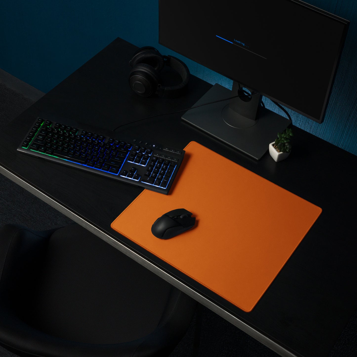 An 18" x 16" orange gaming desk pad sitting on a black desk with a monitor, keyboard, and mouse.