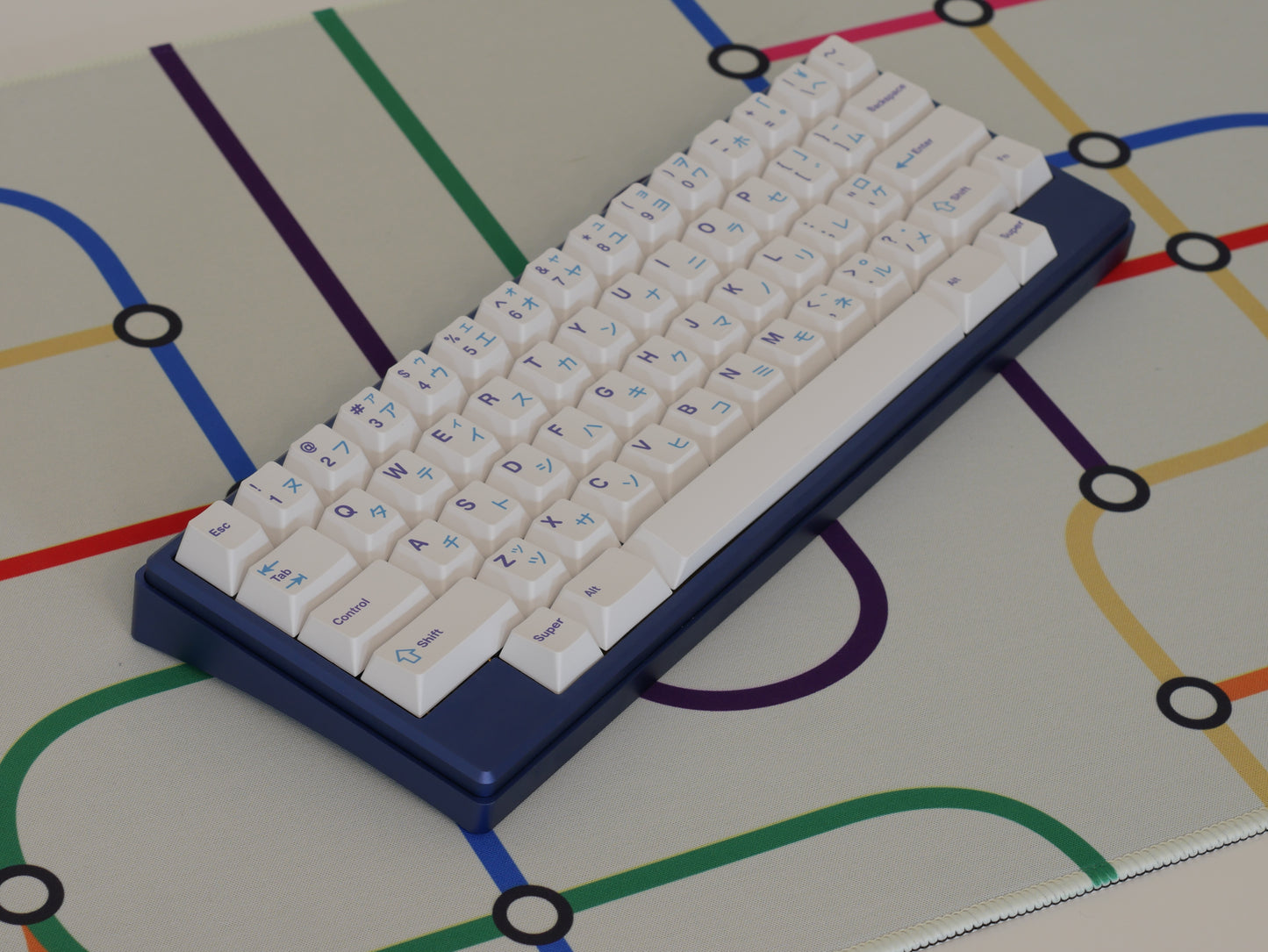 A blue keyboard with white keycaps sits in the middle of the subway transit desk mat with a white background and colorful lines.