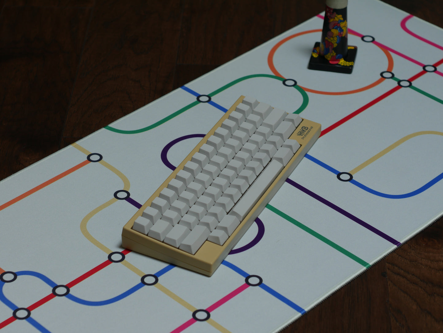 The subway transit desk mat with a white background and colorful lines. A beige keyboard with white keycaps sits on the desk mat.