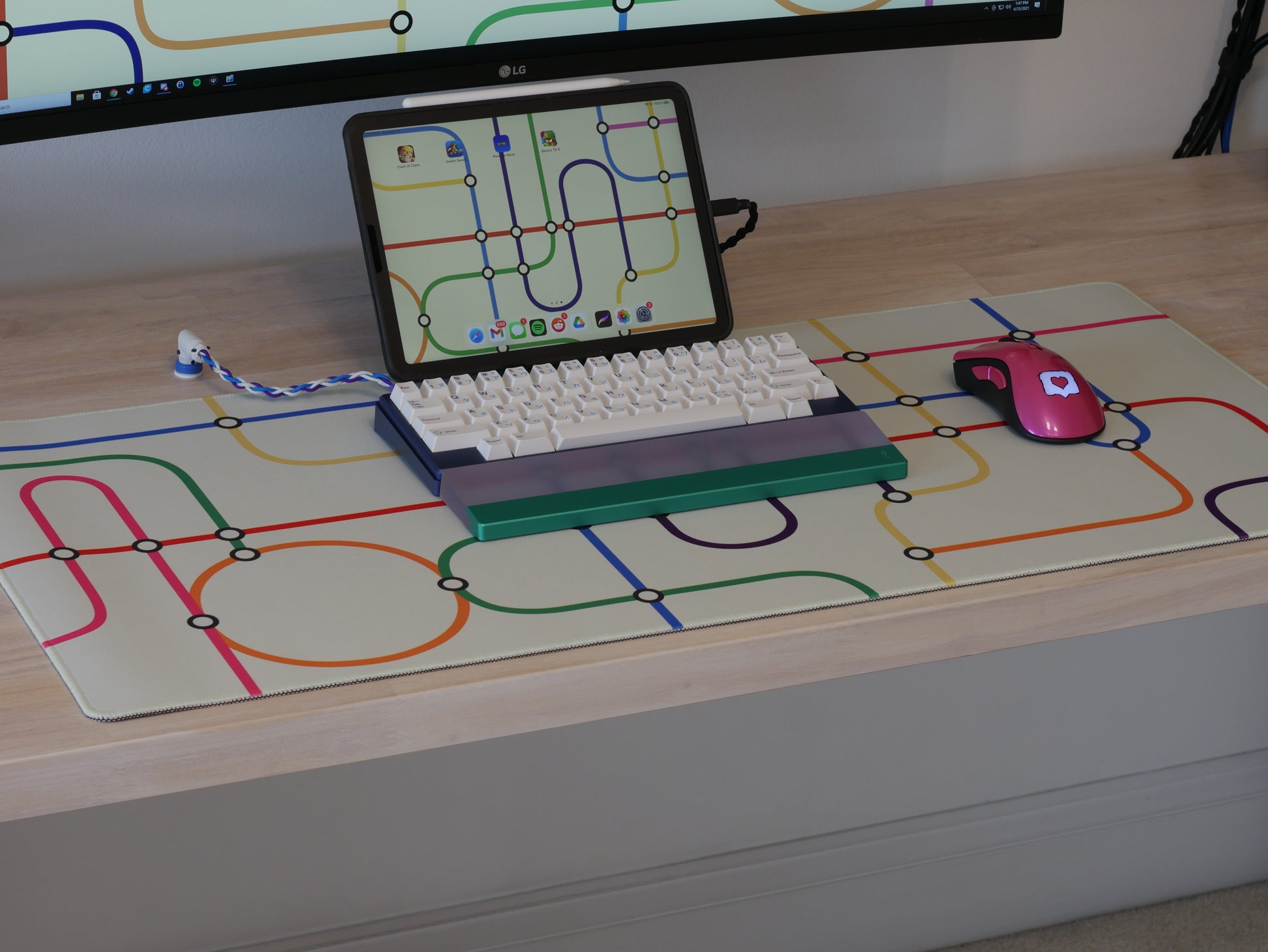 The subway transit desk mat with a white background and colorful lines sitting on a wooden desk. A keyboard, wrist rest, and mouse sit on the desk mat.