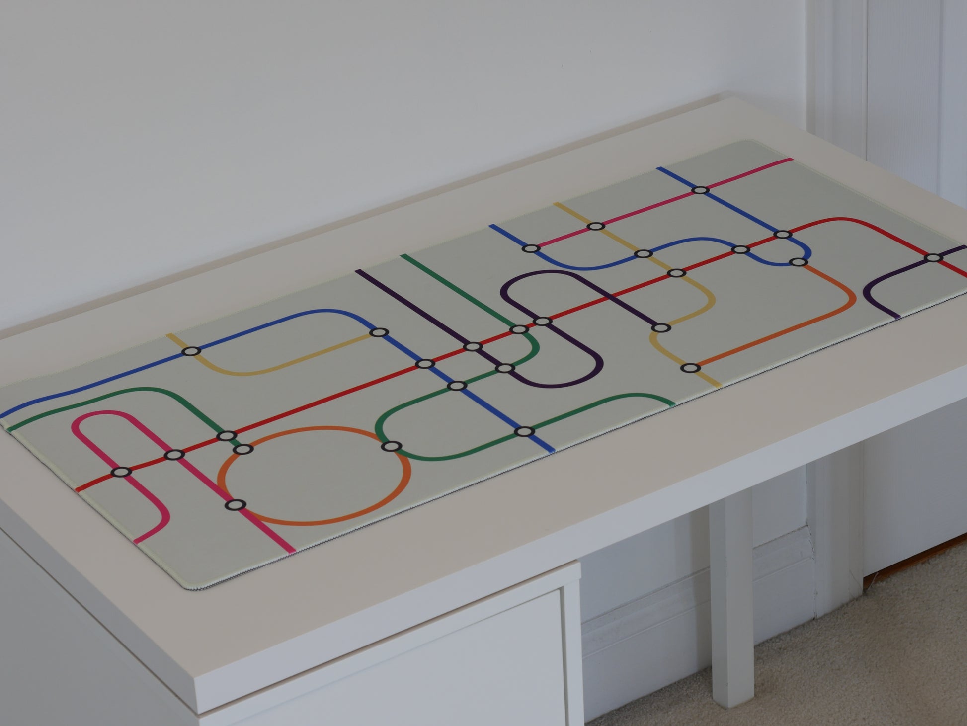 A subway transit desk mat with a white background and colorful lines. The desk mat sits on a white desk.