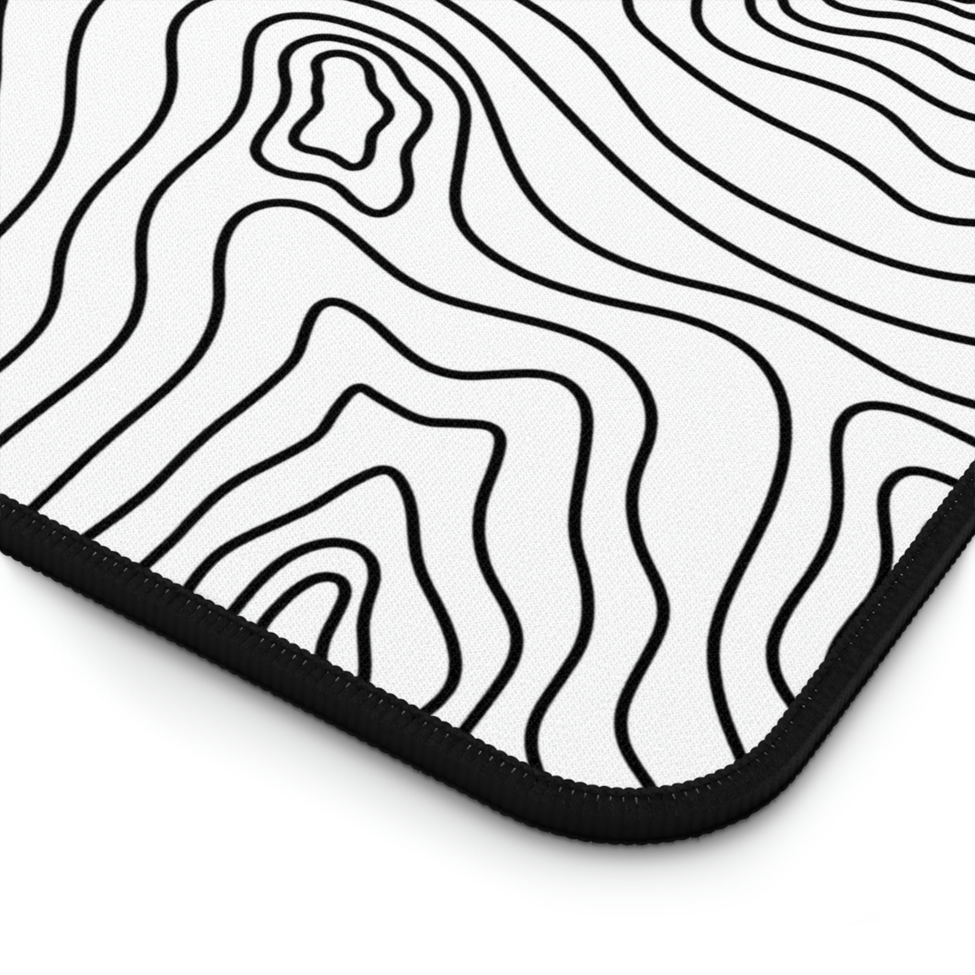 The bottom right corner of a 12" x 18" white desk mat with black topographic lines.