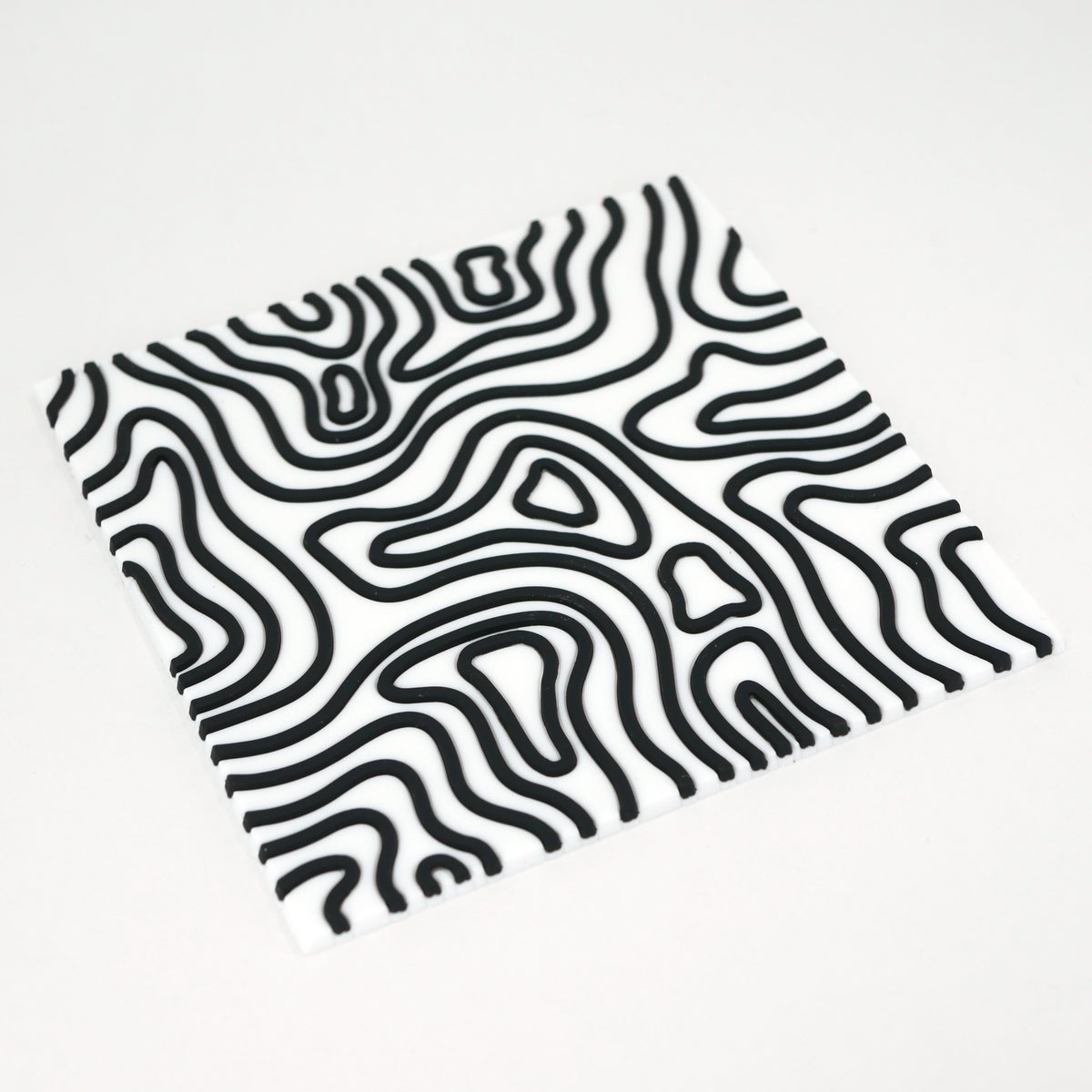 A square white topographic PVC rubber coaster with black lines sitting at an angle.