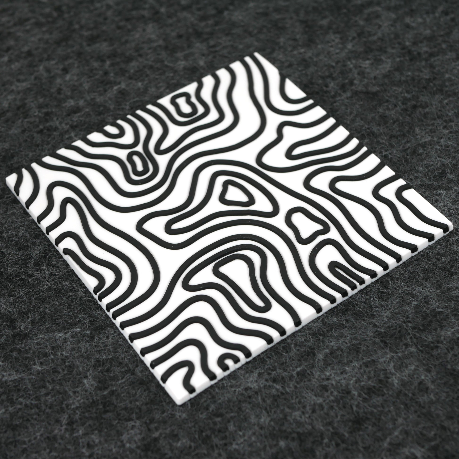 A square white topographic PVC rubber coaster sitting at an angle on a gray desk mat.