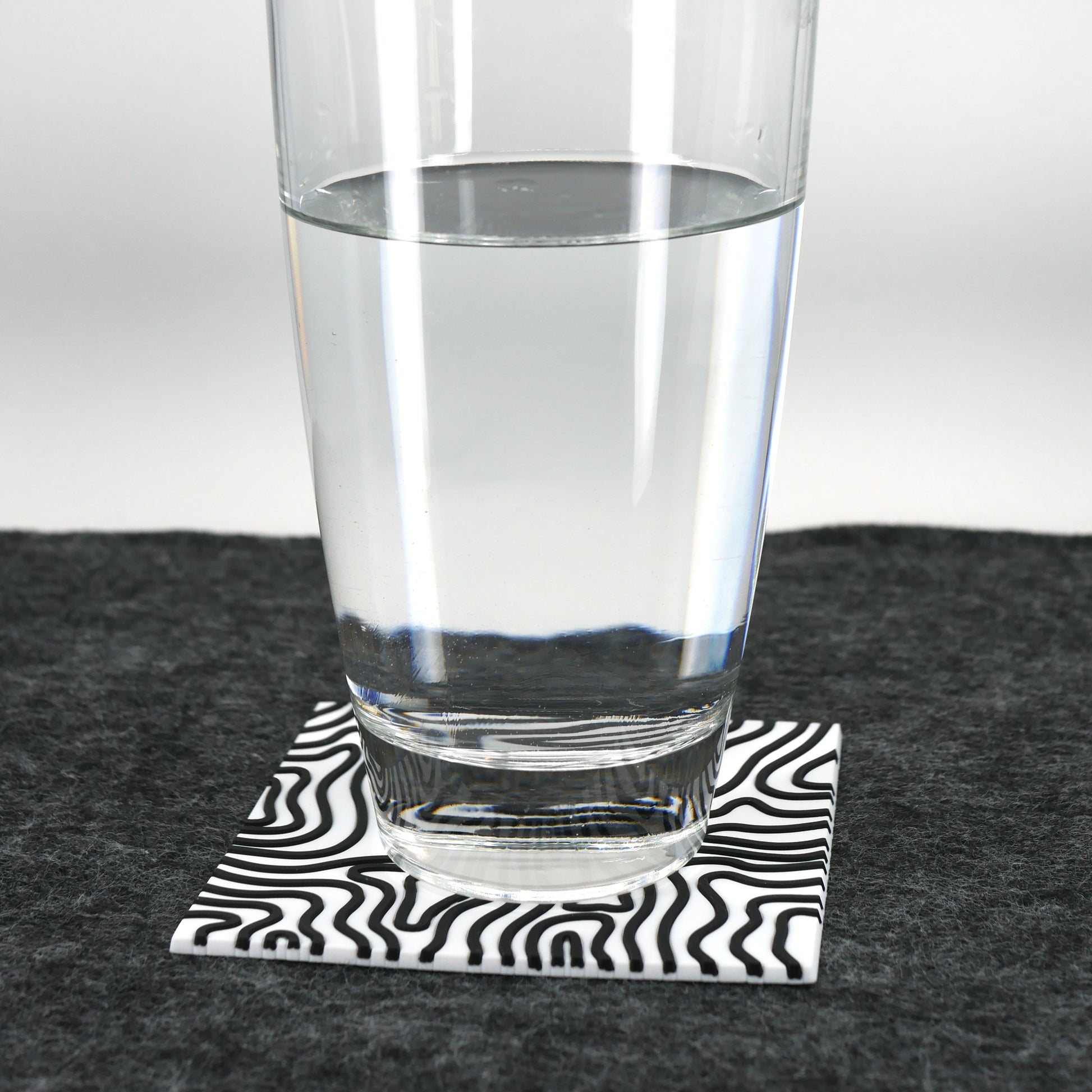 A square white topographic PVC rubber coaster sitting on a gray desk mat. A glass of water sits on the coaster.