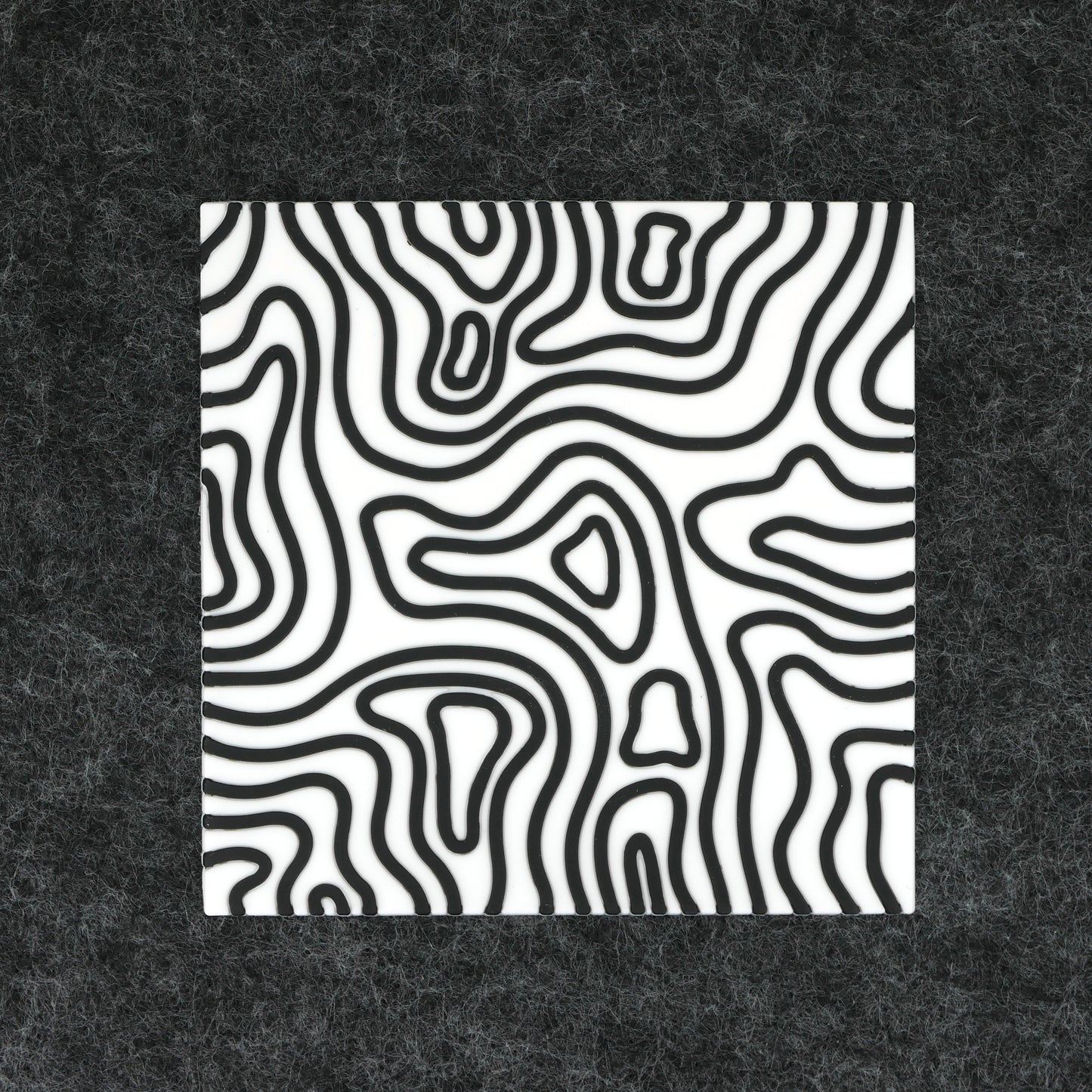 A square white topographic PVC rubber coaster sitting on a gray desk mat.