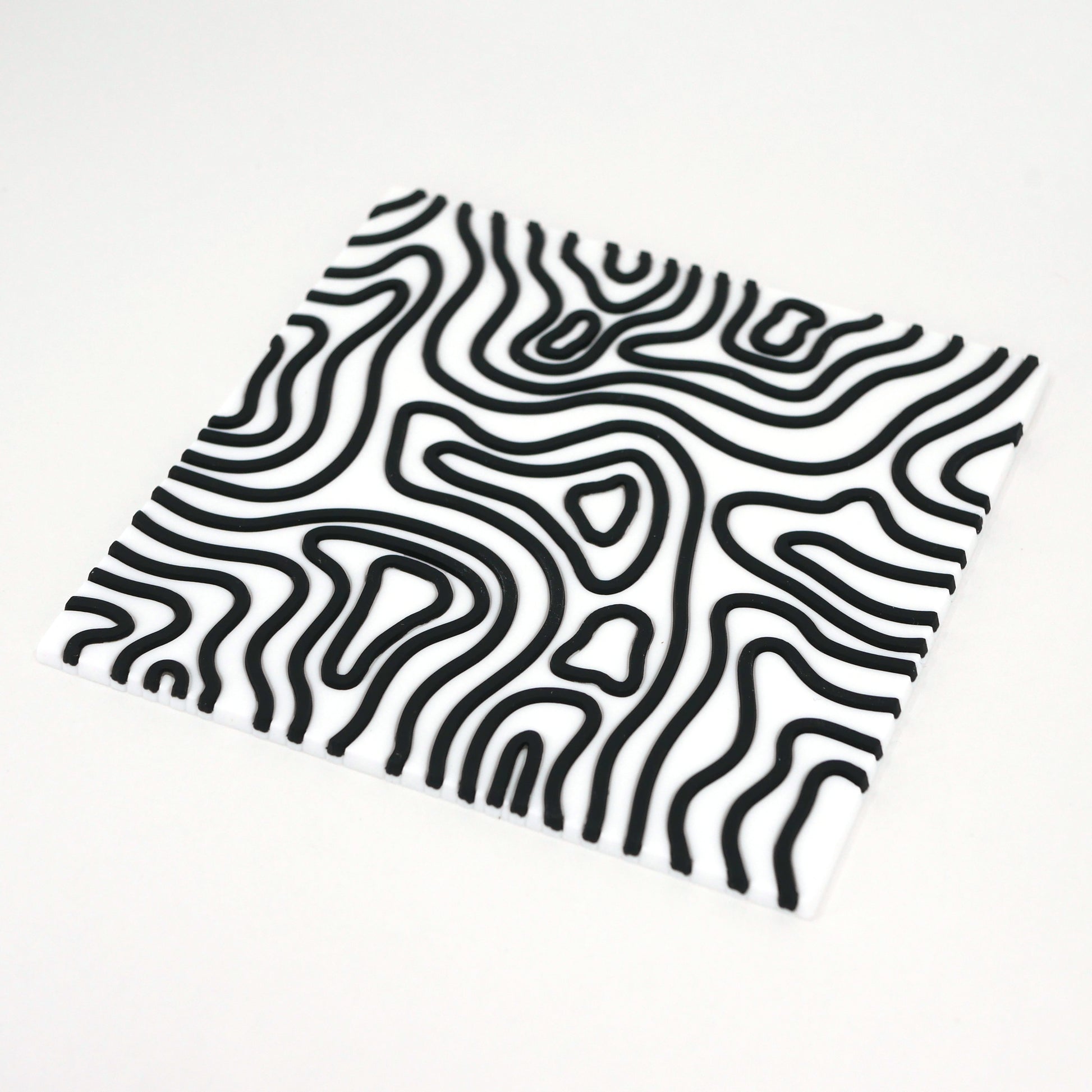 A square white topographic PVC rubber coaster with black lines sitting at an angle.