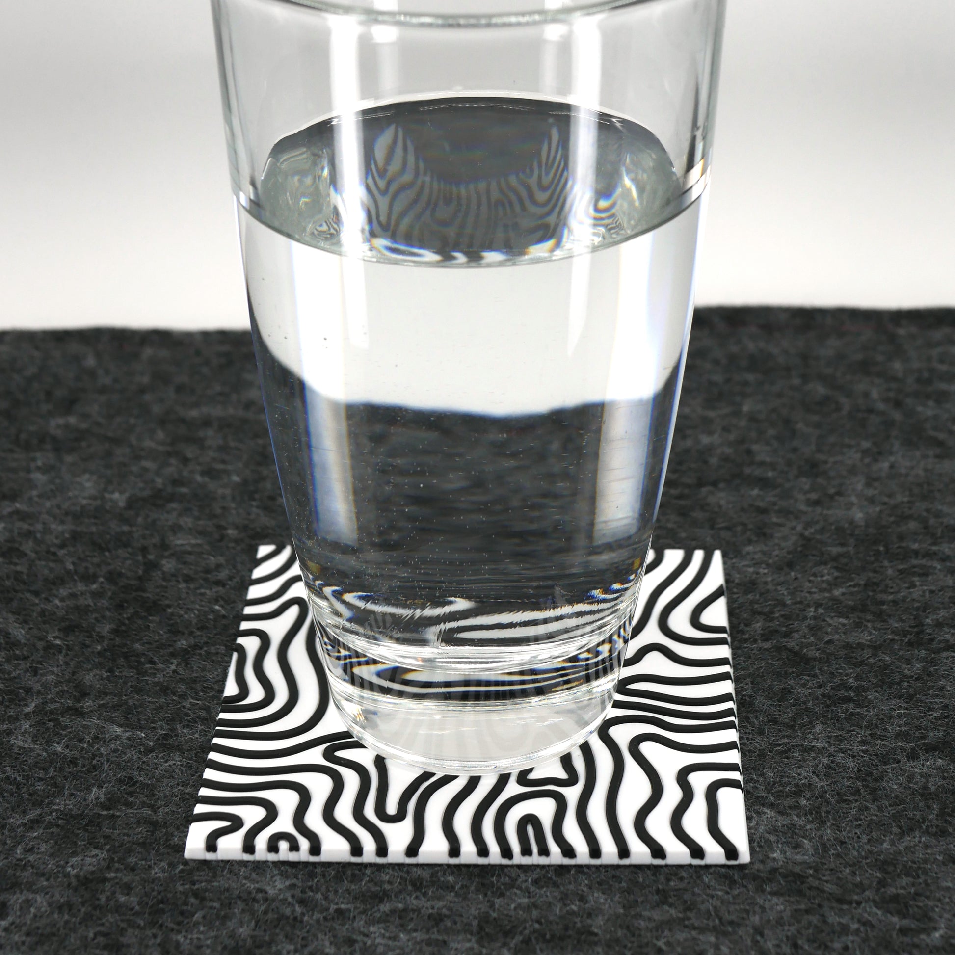A square white topographic PVC rubber coaster sitting on a gray desk mat. A glass of water sits on the coaster.